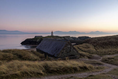 UK, Wales, Newborough, Cottage an der Küste von Ynys Llanddwyn bei Sonnenuntergang mit Twr Mawr-Leuchtturm im Hintergrund - WPEF05940