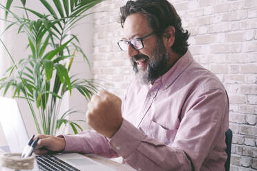 Fröhlicher Geschäftsmann mit gestikulierender Faust, der mit einem Laptop zu Hause sitzt - SIPF02852