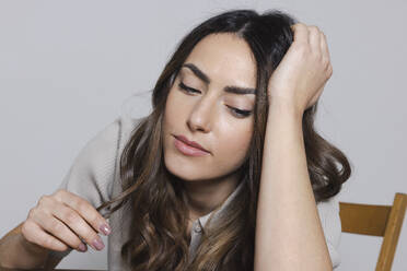 Thoughtful woman with hand in hair sitting on chair against gray background - EIF03848