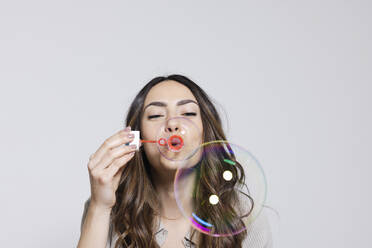 Young woman blowing soap bubbles against gray background - EIF03853