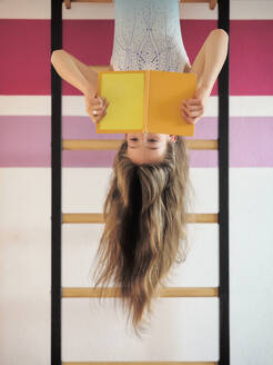 Girl reading book hanging upside down on wall bars - DIKF00683