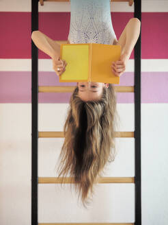 Mädchen liest Buch, das kopfüber an einer Sprossenwand hängt - DIKF00683