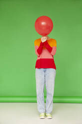 Junge Frau, die ihr Gesicht mit einem roten Luftballon bedeckt, steht vor einem grünen Hintergrund - IYNF00138
