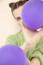 Woman covering face with purple balloon against peach background - IYNF00155