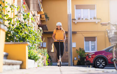 Ganzer Körper einer fröhlichen, molligen Afroamerikanerin mit Schutzhelm, die einen elektrischen Tretroller auf dem Bürgersteig an einem Wohnhaus vorbei an einem sonnigen Tag fährt - ADSF34437