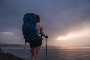 Rückenansicht einer nicht erkennbaren Wanderin mit Rucksack und Trekkingstöcken, die an der Küste in der Nähe des plätschernden Meeres bei Sonnenuntergang steht - ADSF34438
