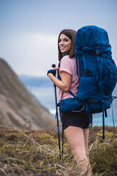 Side view of happy female hiker with backpack and trekking poles looking at camera while standing on coast near rippling sea at sundown time - ADSF34440