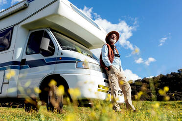 Full body of senior male tourist in hat standing near RV car parked on grassy field while observing nature on summer day - ADSF34441