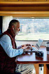 Side view of mature bearded male in wireless earphones typing on netbook while sitting in RV car during road trip - ADSF34443