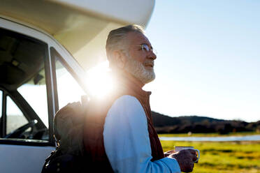 Side view senior male traveler with cup of coffee standing near RV car during road trip in nature - ADSF34449