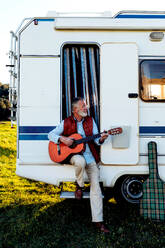 Talented senior bearded male traveler playing song on acoustic guitar while sitting in doorway of RV car during road trip in nature - ADSF34450