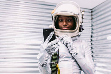 Fokussierte afroamerikanische Frau in weißem Raumanzug mit Helm surft auf einem Mobiltelefon, während sie in einem modernen Raumschiff auf hellem Hintergrund steht - ADSF34456