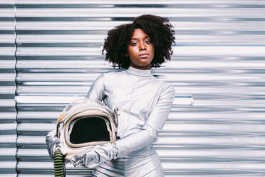 Charming African American female with helmet in white astronaut costume looking at camera while standing in white spaceship on light background - ADSF34457