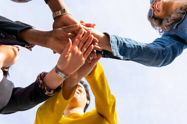 From below group of optimistic women stacking hands together with crop faceless friends while standing on street against blue sky - ADSF34463