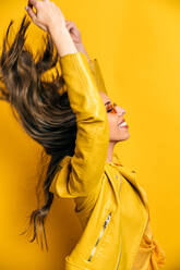 Side view of happy Hispanic female model in trendy sunglasses and bright leather jacket with flowing hair closing eyes and enjoying wind in studio against yellow background - ADSF34473