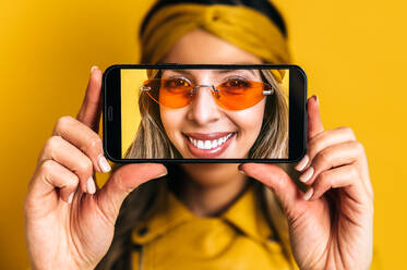 Cheerful female in orange sunglasses and bright leather jacket showing selfie on cellphone in studio on yellow background - ADSF34475