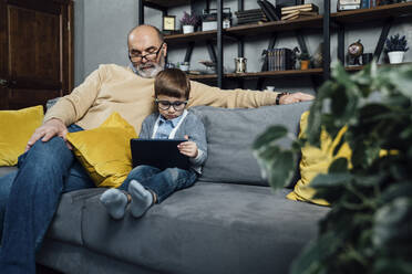 Boy wearing eyeglasses studying through tablet PC sitting by grandfather on sofa at home - VPIF05630