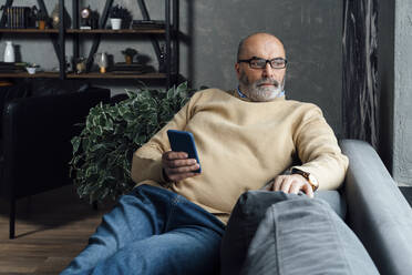 Thoughtful senior man with mobile phone sitting on sofa at home - VPIF05660