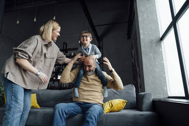 Cheerful woman looking at senior man carrying boy on shoulders in living room at home - VPIF05663