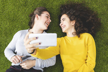 Cheerful friends taking selfie through smart phone lying down on grass at park - WPEF05948