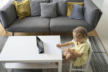 Girl drawing on paper with pencil sitting on chair at table in living room - SVKF00080