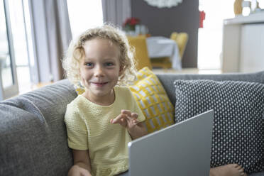 Glückliches blondes Mädchen mit Laptop auf dem Sofa im Wohnzimmer zu Hause - SVKF00085