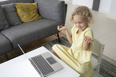 Blondes Mädchen sitzt gestikulierend mit Laptop am Tisch im Wohnzimmer - SVKF00088