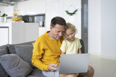 Father sharing laptop with daughter sitting on sofa at home - SVKF00090