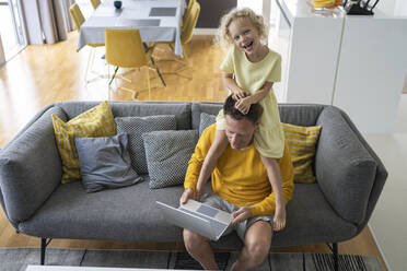 Playful daughter sitting on shoulder of father using laptop at home - SVKF00093