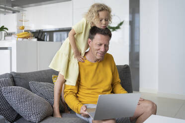 Cute daughter leaning on father sitting with laptop on sofa at home - SVKF00096