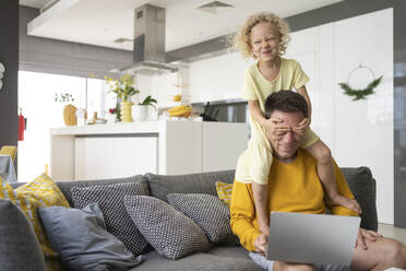 Die schelmische Tochter verdeckt die Augen des Vaters, der zu Hause auf dem Sofa mit dem Laptop sitzt - SVKF00097