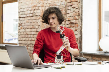 Smiling teenage boy with robotic arm using laptop sitting at table - DHEF00647