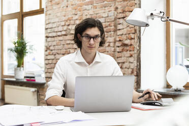 Geschäftsmann mit Laptop sitzt am Schreibtisch im Büro - DHEF00648