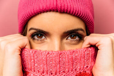 Attractive female in hat covering half of face with knitted sweater while looking at camera on pink background in studio - ADSF34481