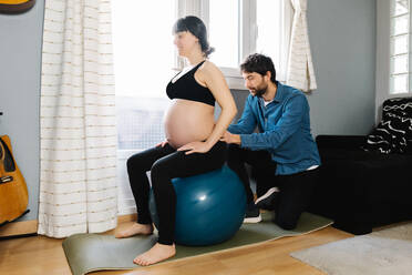 Side view of loving husband helping pregnant wife doing exercise on fit ball during training at home - ADSF34494