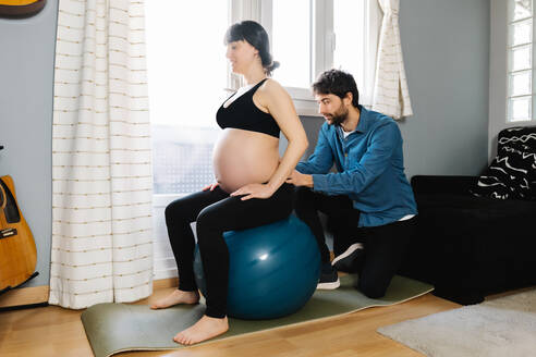 Seitenansicht des liebenden Ehemannes, der seiner schwangeren Frau bei der Übung auf dem Fit-Ball hilft, während des Trainings zu Hause - ADSF34494