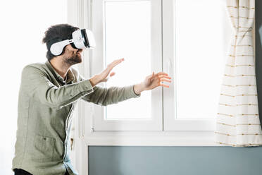 Young bearded male with dark hair in casual clothes gesturing and smiling while experiencing virtual reality in modern headset standing near window at home - ADSF34501