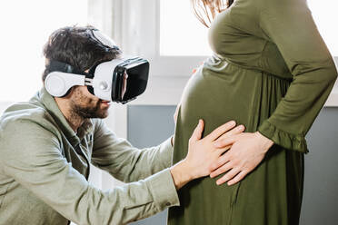 Seitenansicht eines aufmerksamen jungen ethnischen bärtigen Mannes in Freizeitkleidung, der den Bauch seiner unerkennbaren schwangeren Frau berührt, während er zu Hause mit einer modernen VR-Brille virtuelle Realität erlebt - ADSF34502