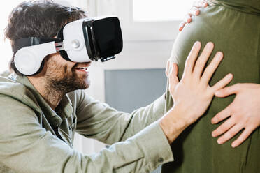 Seitenansicht eines aufmerksamen jungen ethnischen bärtigen Mannes in Freizeitkleidung, der den Bauch seiner unerkennbaren schwangeren Frau berührt, während er zu Hause mit einer modernen VR-Brille virtuelle Realität erlebt - ADSF34503