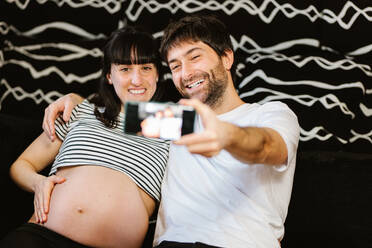 Selective focus of cellphone in hands of smiling pregnant woman showing bare tummy and hugging husband while taking self portrait - ADSF34506