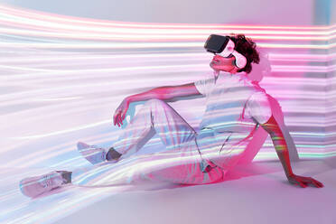 Ganzer Körper eines Mannes in einer modernen VR-Brille auf weißem Hintergrund im Studio mit leuchtenden Lichtern sitzend - ADSF34521