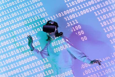 Frau in Freizeitkleidung und moderner schwarzer VR-Brille mit verschiedenen projizierten Zahlen beim Erkunden des Cyberspace in einem Raum - ADSF34523