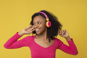 Smiling young woman listening music through wireless headphones against yellow back ground - EIF03899