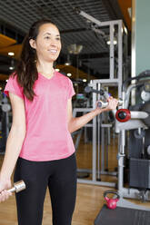 Lächelnde junge Frau beim Training mit Hanteln im Fitnessstudio - IFRF01571
