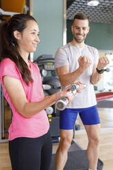 Smiling woman practicing with dumbbells by instructor in gym - IFRF01574