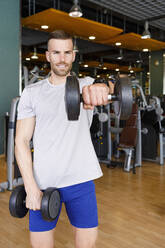 Lächelnder junger Mann beim Training mit Hanteln im Fitnessstudio - IFRF01584