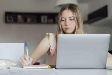 Junge Frau schreibt auf einem Notizblock und sitzt mit einem Laptop zu Hause - PGF01065