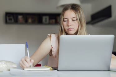 Young woman writing on note pad sitting with laptop at home - PGF01065