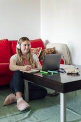 Young woman wearing headphones looking at laptop sitting in living room - PGF01068