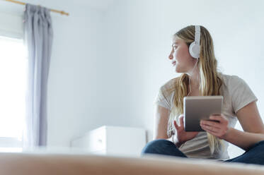 Woman listening music through headphones sitting with tablet PC on bed at home - PGF01073